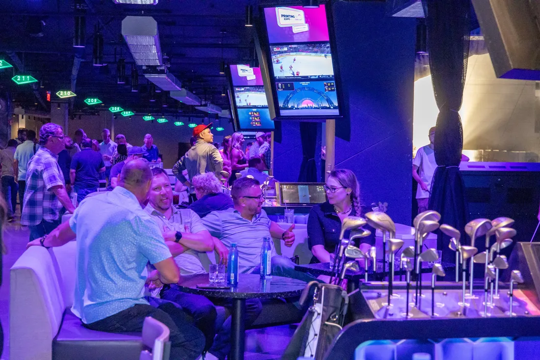 Trade show attendees sitting and talking around tables in a bought-out entertainment venue, with golf bays and event screens behind them.