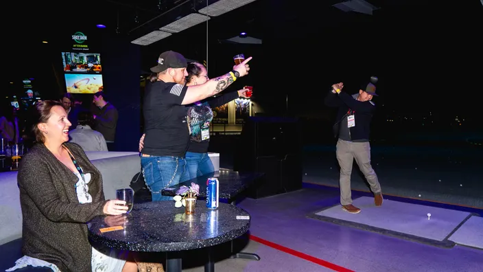 Guests cheering from a table beside a golf bay while another takes a swing.